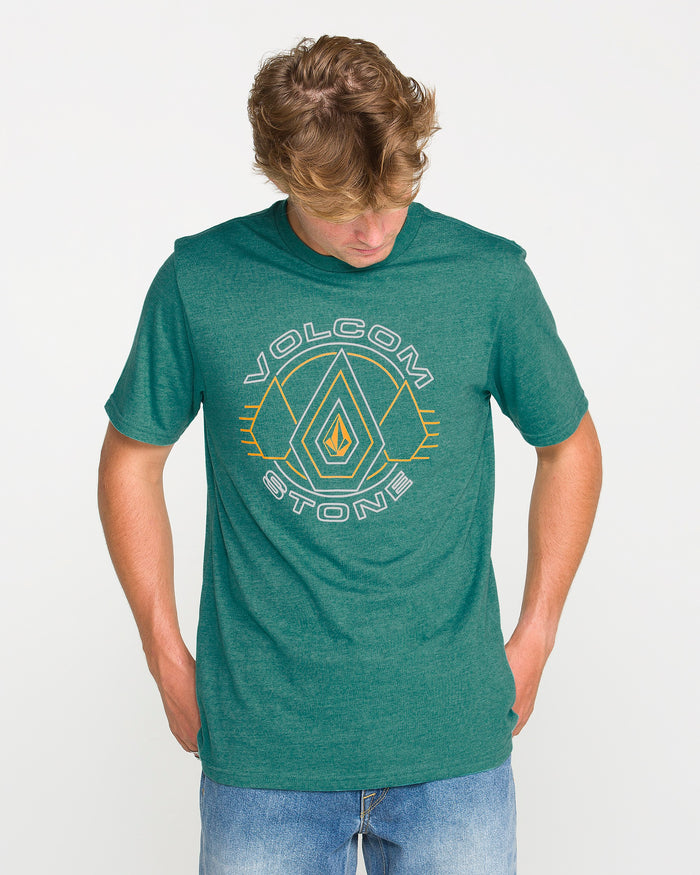 Wearing the Volcom Men's Line Feld Short Sleeve Tee in Miami Green, which features a Volcom Stone soft-hand screen print, and blue jeans, a person stands against a plain white background looking down with hands in their pockets.