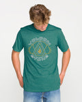 Wearing the Volcom Men's Line Feld Short Sleeve Tee in Miami Green, which features a Volcom Stone soft-hand screen print, and blue jeans, a person stands against a plain white background looking down with hands in their pockets.