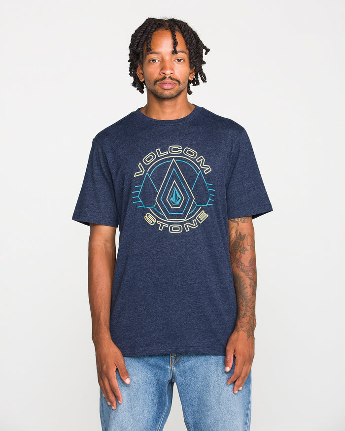 A person wearing a Volcom Men's Line Feld Short Sleeve Tee in navy blue heather, made from soft ring spun cotton, and light blue jeans stands against a plain white background, looking at the camera with a neutral expression.