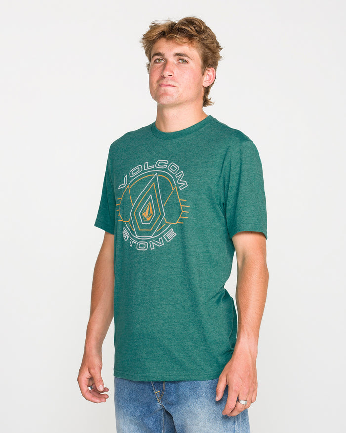 A person wearing the Volcom Men's Line Feld Short Sleeve Tee in Miami Green, featuring a soft-hand screen print, with blue jeans stands against a plain white background, looking slightly to the side with a neutral expression.