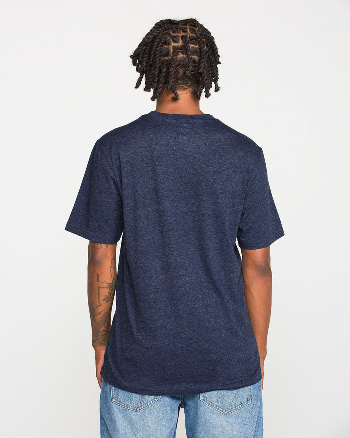 A person with short twists is standing facing away from the camera against a plain white background, wearing the Volcom Men's Line Feld Short Sleeve Tee in dark blue and light blue jeans.