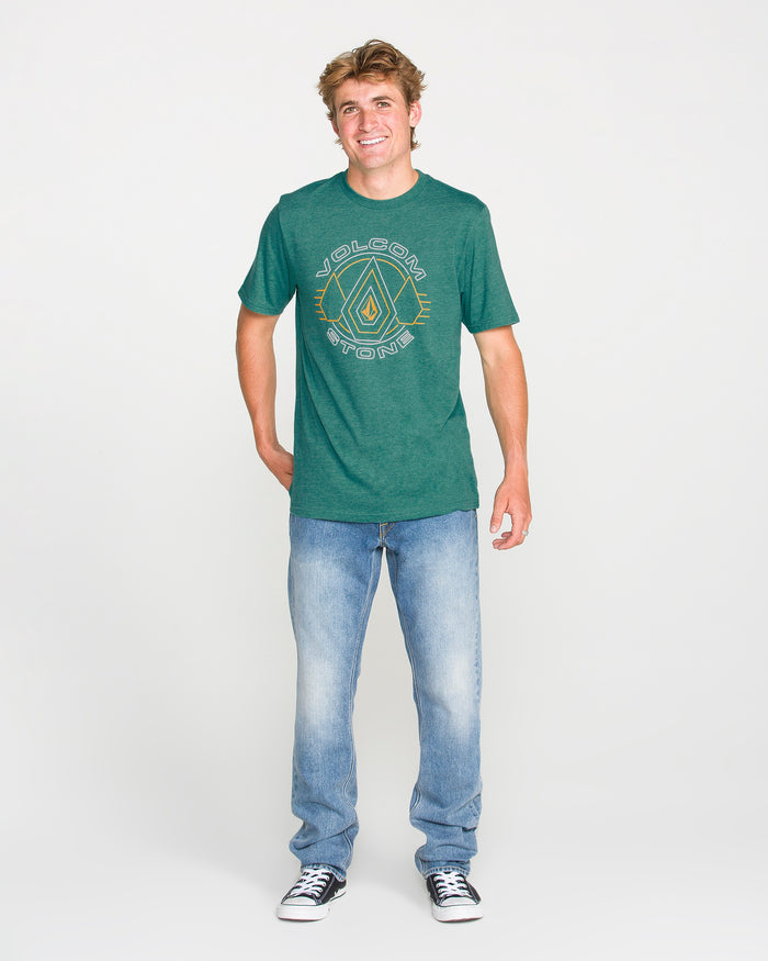 A smiling person stands facing forward, wearing the Volcom Men's Line Feld Short Sleeve Tee in Miami Green, featuring a soft-hand screen print, paired with light blue jeans and black sneakers, against a plain white background.