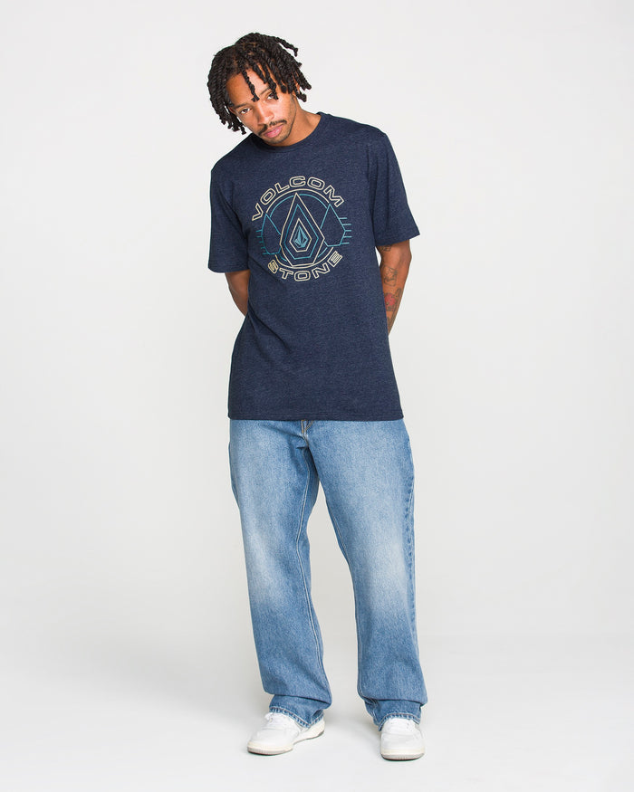 Man with short dreadlocks wears a navy Volcom Men's Line Feld Short Sleeve Tee, crafted from soft ring spun cotton, paired with light blue jeans and white sneakers, standing against a plain white background.