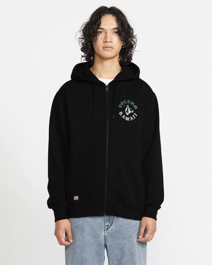 A person with wavy dark hair models the Volcom Men's Lineage Zip Fleece, a relaxed fit black hoodie with a small green and white left chest graphic, paired with light blue jeans against a plain white background.