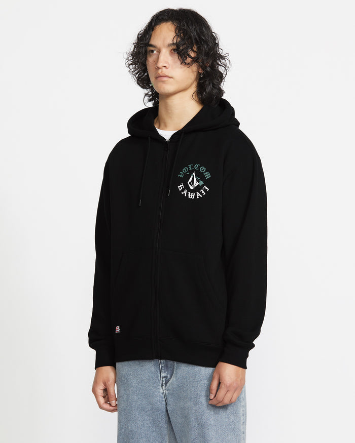 A person with curly hair wears the Volcom Men's Lineage Zip Fleece, a relaxed fit black hoodie with a small graphic and text on the left chest, paired with light blue jeans, standing against a plain white background.