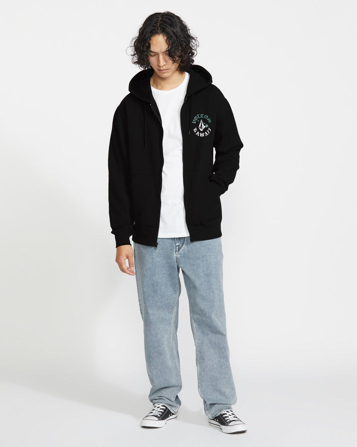 A person with curly hair wears the Volcom Men's Lineage Zip Fleece hoodie in black, paired with a white tee, light blue jeans, and black-and-white sneakers, standing against a white background with hands in pockets.