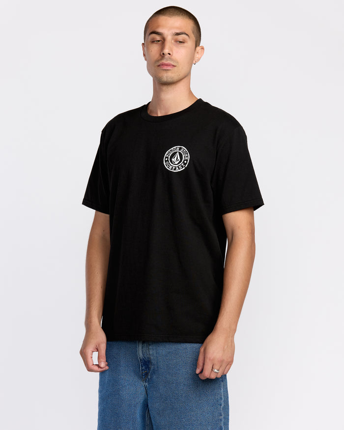 A person wearing the Volcom Men’s Logo Stamp Short Sleeve Tee, crafted from premium ring-spun cotton with a small white logo on the left chest, paired with blue jeans, stands against a plain white background.