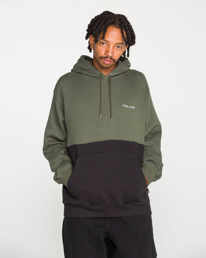 A person stands with hands in pockets, looking at the camera while wearing the Volcom Men's Long Division Pullover—a green and black colorblocked hoodie made with REPREVE recycled fibers and featuring "VOLCOM" on the chest. The background is plain white.
