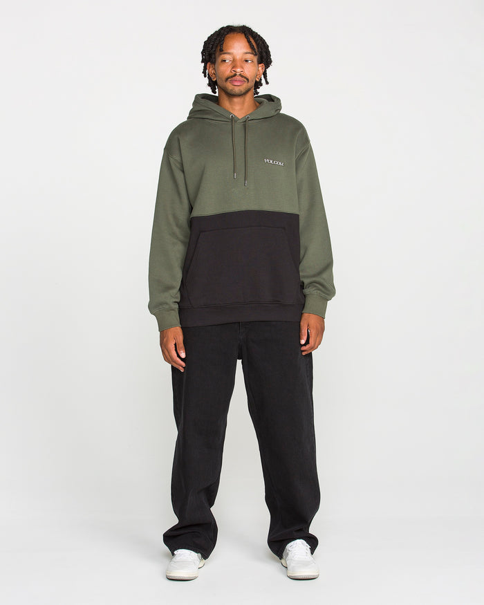 A person stands facing forward, wearing the Volcom Men's Long Division Pullover—a green and black colorblocked hoodie made with REPREVE recycled fibers—paired with black pants and white sneakers, against a plain white background.