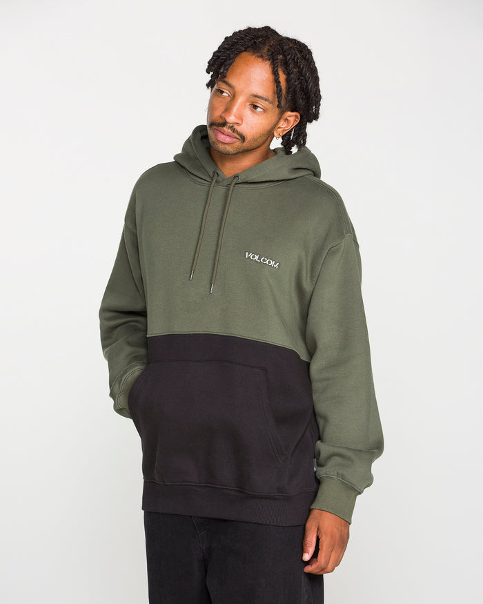 A man stands against a plain white background, looking to his left with one hand in his pocket, wearing the Volcom Men's Long Division Pullover. This olive green and black colorblocked hoodie features "VOLCOM" emblazoned on the chest and is crafted using REPREVE recycled fibers for an eco-conscious take on contemporary style.
