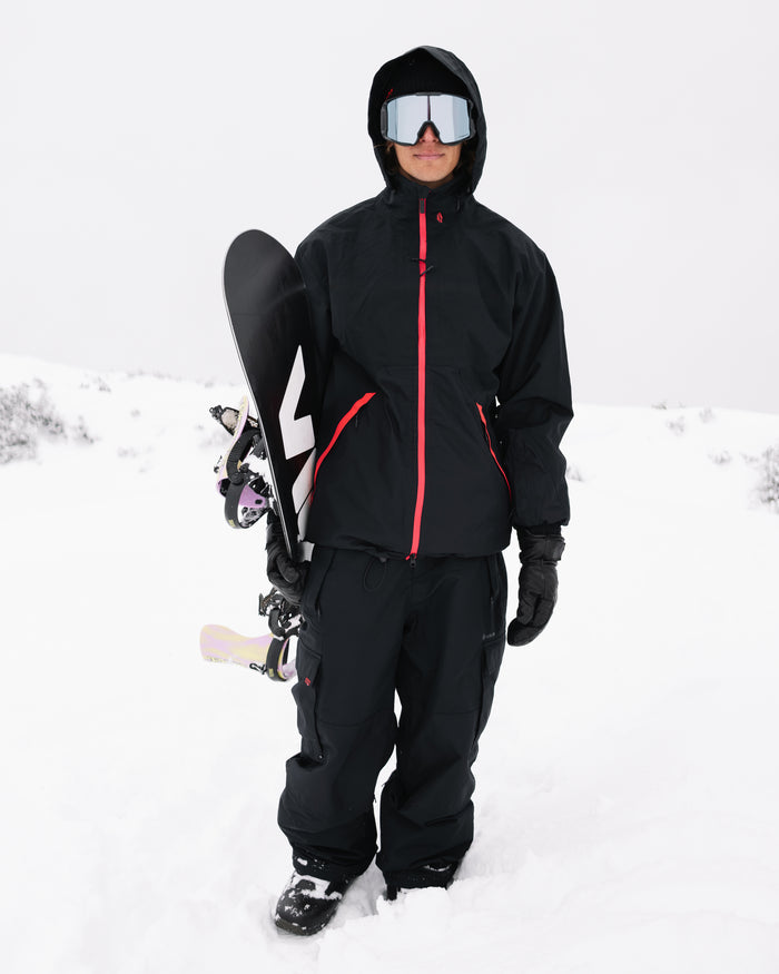 A person wearing the Volcom Men’s Longo GORE-TEX Jacket along with snow goggles and gloves stands in the snow, holding a snowboard upright on their left side against an overcast, snowy background.