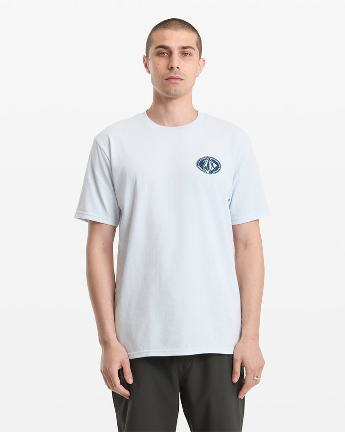 A person with short hair, wearing the Volcom Men's Loopy Short Sleeve Tee in light blue with a small dark chest graphic and dark pants, stands against a plain white background.