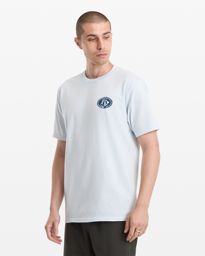 A man with short hair models the Volcom Men's Loopy Short Sleeve Tee in light blue, featuring a small circular logo on the left chest, standing against a plain white background and glancing slightly to his left.
