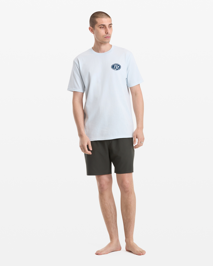 A man with short hair stands barefoot against a white background, wearing Volcom's Men's Loopy Short Sleeve Tee in light blue with a small circular chest logo, paired with black shorts.