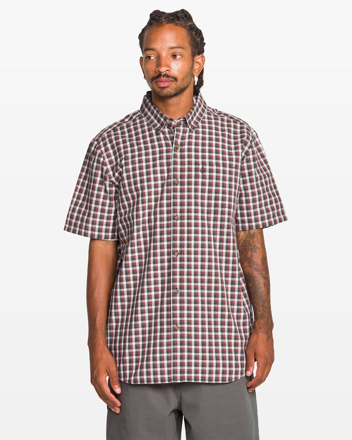 A man with long braided hair and a tattooed right arm wears a Volcom Men's Louie Check Short Sleeve Tee in light blue acid check with gray pants, standing against a plain white background.
