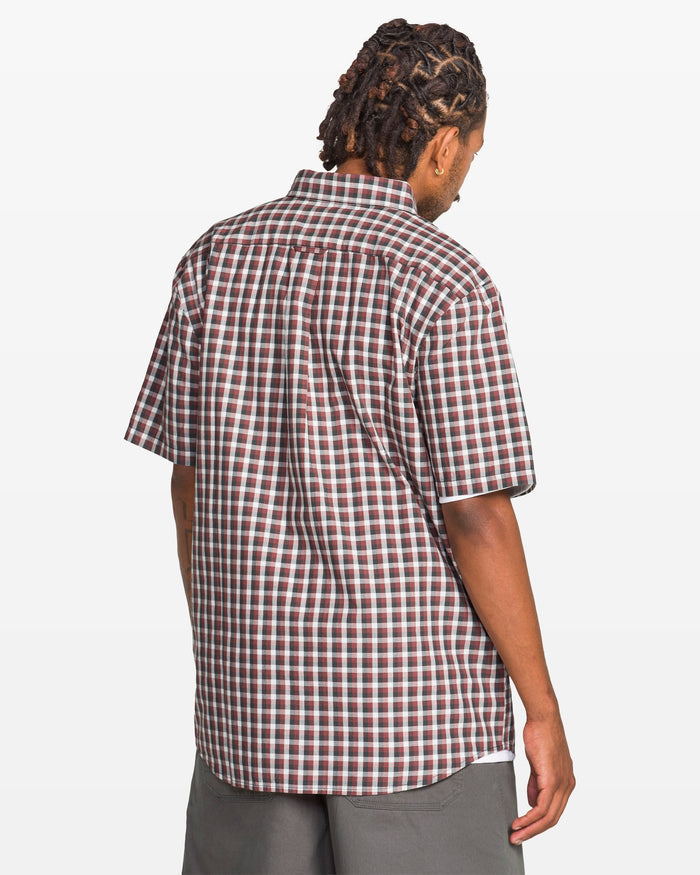 A man with short dreadlocks is shown from the back, wearing a Volcom Men's Louie Check Short Sleeve Tee in a red, white, and black checkered pattern with gray shorts against a plain white background.