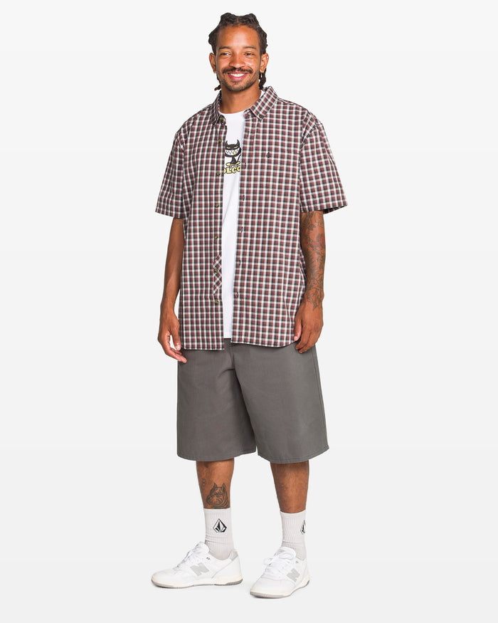 A man smiles in a Volcom Men's Louie Check Short Sleeve Tee in light blue acid check over a white graphic T-shirt, paired with gray knee-length shorts, white crew socks, and white sneakers against a plain light background.