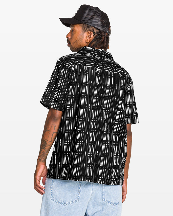 Man wearing the Volcom Men's Macros Short Sleeve Tee, featuring a black allover print and boxy fit, paired with light blue jeans and a black cap, facing away over his shoulder against a plain white background.