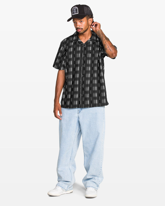 A man wearing the Volcom Men's Macros Short Sleeve Tee, light blue baggy jeans, white sneakers, and a black baseball cap stands against a plain white background, touching the back of his neck with one hand.