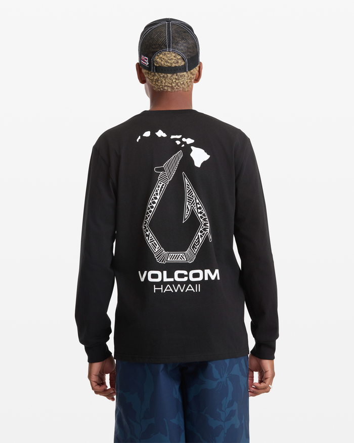 A man is seen from behind wearing the Volcom Men's Makau Long Sleeve Tee with a shark and Hawaiian islands graphic and "VOLCOM HAWAII" text on the back, paired with a mesh cap and blue camo pants.
