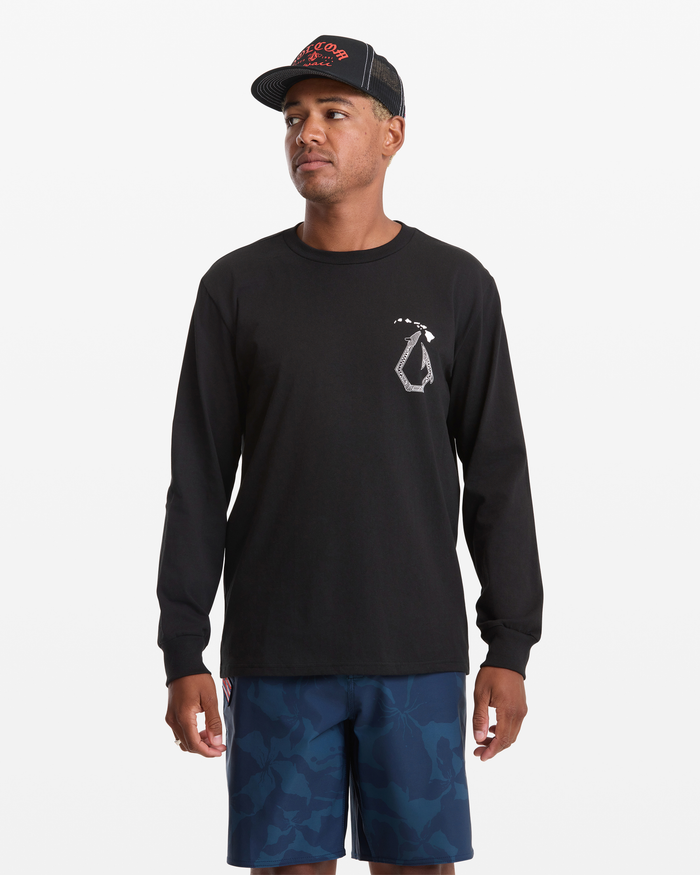 A man wears the Volcom Men's Makau Long Sleeve Tee in black, featuring a small white chest logo, paired with blue patterned shorts and a black and red cap, standing against a plain white background.