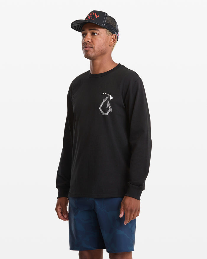 A man wears the Volcom Men's Makau Long Sleeve Tee in black with a white chest graphic, paired with blue camo shorts and a black mesh cap, standing against a plain white background.