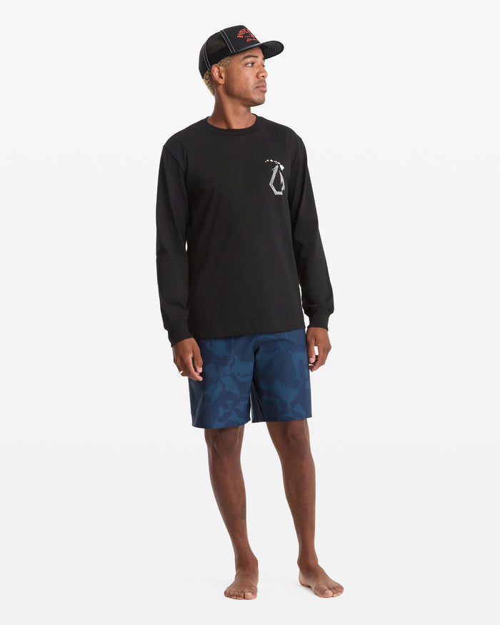 A man stands barefoot against a plain white background, wearing the Volcom Men's Makau Long Sleeve Tee in black, paired with blue camo shorts and a black cap.