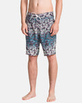 A man stands barefoot in Volcom Men's Manic Boardshorts featuring a tropical leaf pattern in green, purple, and teal with a white drawstring waist. Only his torso and legs are visible.