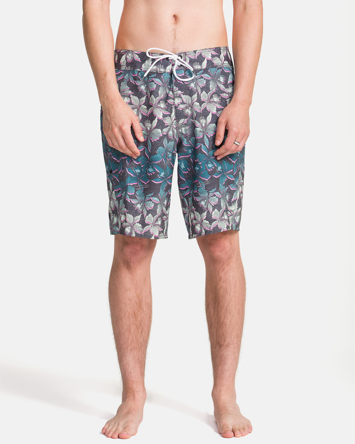 A man stands barefoot in Volcom Men's Manic Boardshorts, featuring a drawstring waist and a tropical leaf pattern in green, blue, and purple on white. The background is plain white.