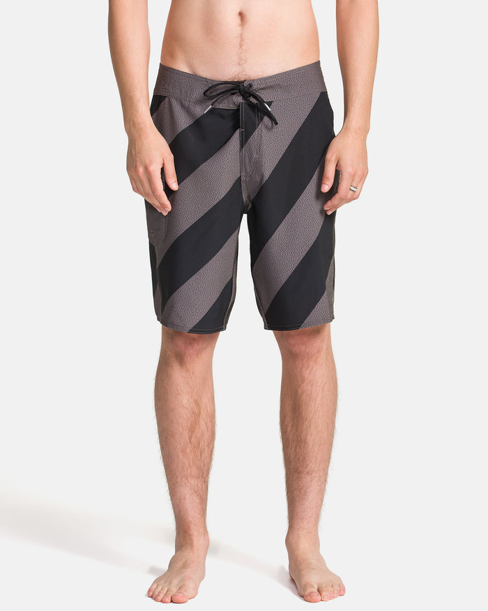 A person stands barefoot in Volcom Men's Manic Boardshorts, featuring knee-length black and gray diagonal stripes, shown from the front against a plain white background.