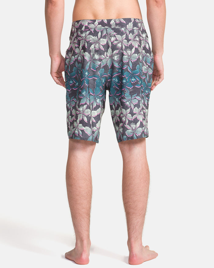 A person stands barefoot facing away, wearing Volcom Men's Manic Boardshorts featuring a purple, teal, and gray tropical leaf pattern on a white background.