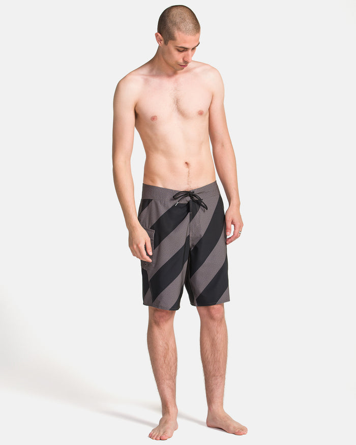 A man with short hair stands barefoot, wearing Volcom Men's Manic Boardshorts—black-and-gray striped and knee-length—with his arms relaxed by his sides against a plain white background.