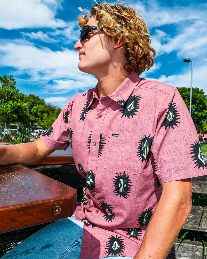 A person with curly blond hair and sunglasses sits outdoors at a wooden table on a sunny day, wearing the Volcom Men's Markerstone Short Sleeve Shirt—a pink shirt with black and green geometric prints. Blue sky and greenery are in the background.
