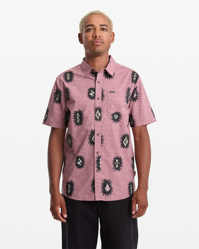 A person with short blond hair wears the Volcom Men's Markerstone Short Sleeve, featuring a black and white abstract sunburst print on pink, standing against a plain white background.