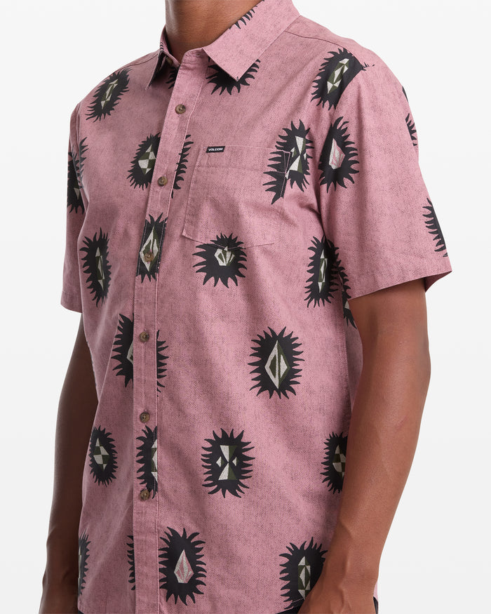 A person wears the Volcom Men's Markerstone Short Sleeve shirt in light maroon with a black geometric print, pointed collar, chest pocket, and small logo tag.