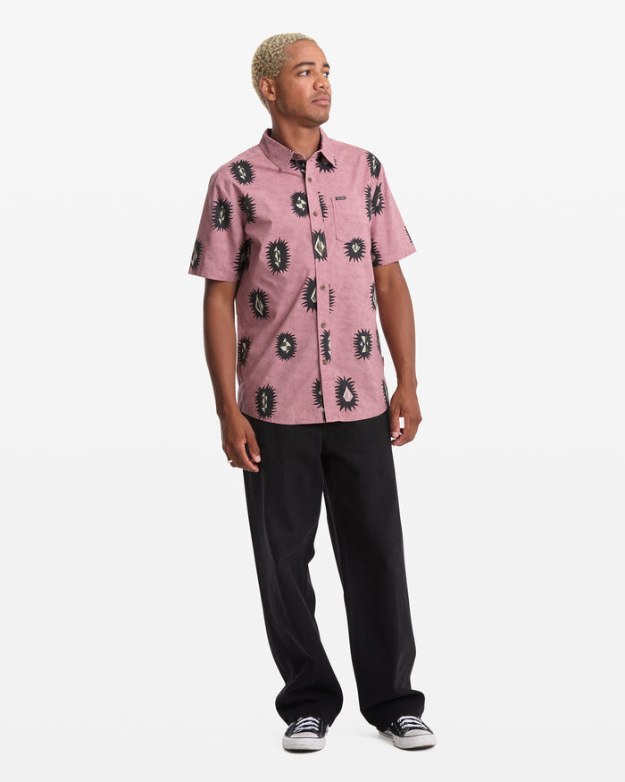 A person with short, light blond hair wears a pink Volcom Men's Markerstone Short Sleeve shirt with black print, black pants, and black sneakers, standing against a plain white background.