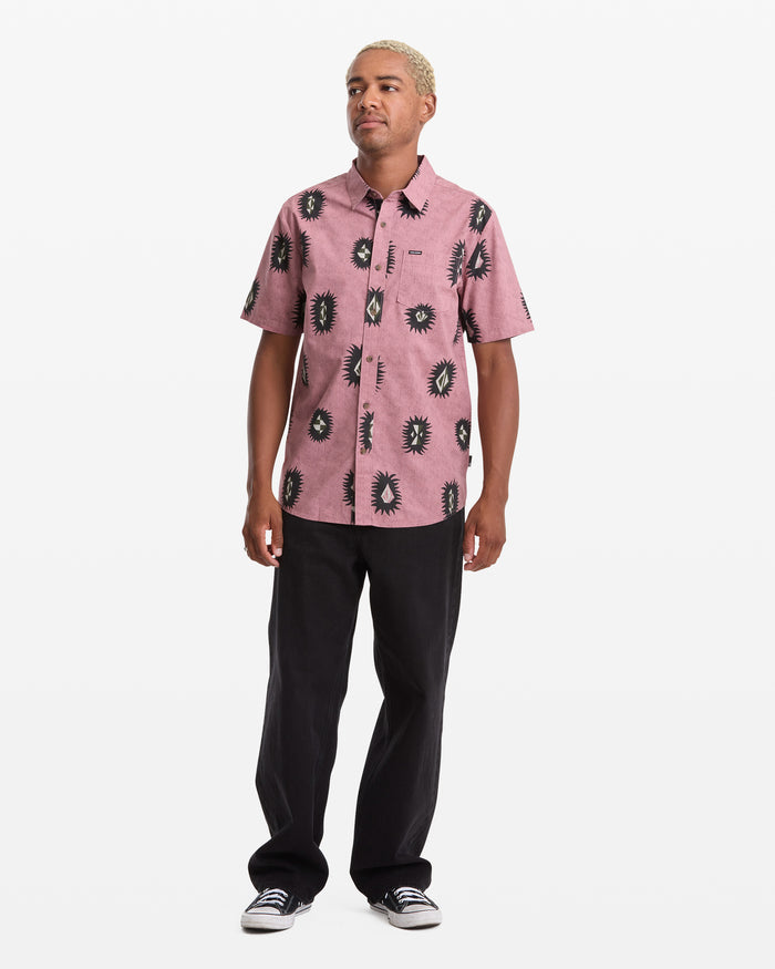 A person stands against a white background, wearing the Volcom Men's Markerstone Short Sleeve shirt in pink with black allover print, paired with black pants and black-and-white sneakers.