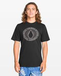 A person with long brown hair wears a Volcom Men's Medalist Short Sleeve Tee with a modern fit and circular geometric graphic, styled with blue shorts featuring a white leaf print. The ring-spun heather fabric adds comfort against a plain background.