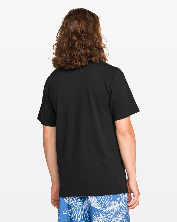 A person with long, curly brown hair is seen from behind in a modern fit plain black Volcom Men's Medalist Short Sleeve Tee and blue shorts with a white tropical leaf pattern.