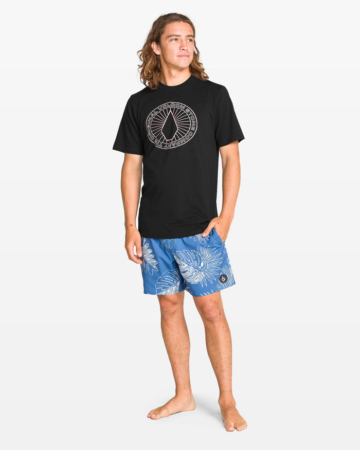 A young man with long hair stands barefoot in a Volcom Men's Medalist Short Sleeve Tee featuring a circular graphic and blue floral shorts, gazing to the side against a plain white background.