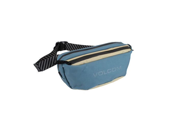 The Volcom Men's Mini Dos Pack Waist Bag, shown in blue and beige with a black adjustable strap and a zippered front pocket, is displayed on a white background.