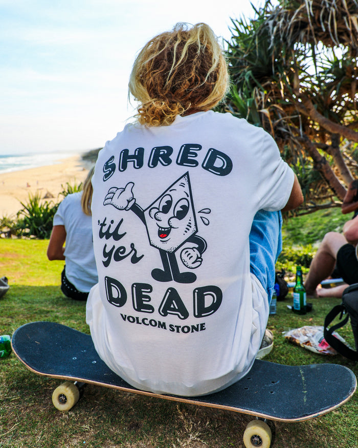 Wearing Volcom's Men's Mister Stone Short Sleeve Tee, a person with long blonde dreadlocks sits on a skateboard at a grassy beachside spot with others nearby.