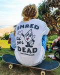Wearing Volcom's Men's Mister Stone Short Sleeve Tee, a person with long blonde dreadlocks sits on a skateboard at a grassy beachside spot with others nearby.