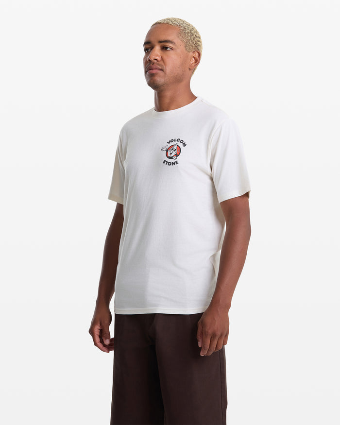 A person with short blond hair wears the Volcom Men's Mister Stone Short Sleeve Tee, featuring a small circular graphic and text on the chest, with dark pants, standing against a plain white background.