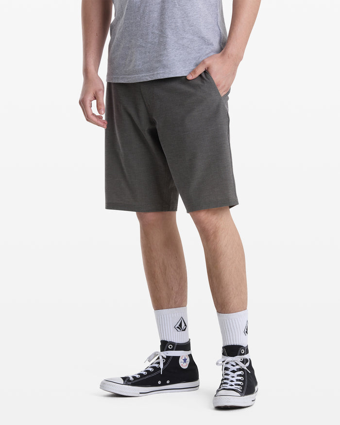 A person wears a gray t-shirt, Volcom Men's Mix Frickin Cross Shred Shorts in dark gray, white crew socks with a black logo, and black Converse high-top sneakers, standing with one hand in their pocket against a plain white background.