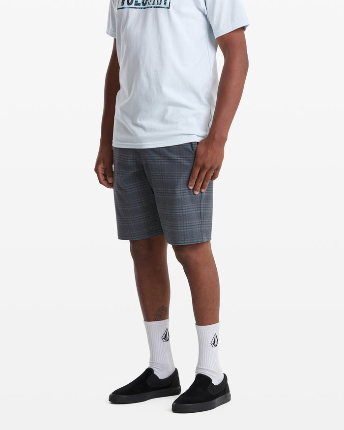 A person stands against a white background wearing a light blue T-shirt, Volcom Men's Mix Frickin Cross Shred Shorts in gray plaid, white socks with a black logo, and black slip-on shoes.