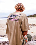 A person with wavy, shoulder-length hair sits on a beach facing away, wearing the Volcom Men's Mod Label Short Sleeve Tee in tan and green shorts. The ocean and a cloudy sky stretch out in the background.