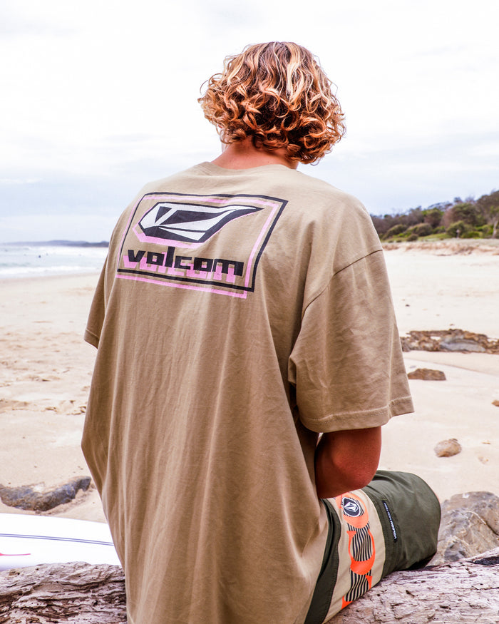 A person with wavy, shoulder-length hair sits on a beach facing away, wearing the Volcom Men's Mod Label Short Sleeve Tee in tan and green shorts. The ocean and a cloudy sky stretch out in the background.