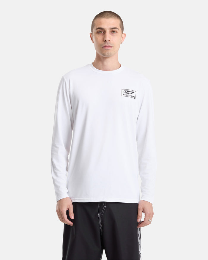 A man wearing a Volcom Men's Modstone Loose Long Sleeve rashguard, featuring UV protection and a small rectangular logo on the left chest, paired with black shorts, stands against a plain white background.