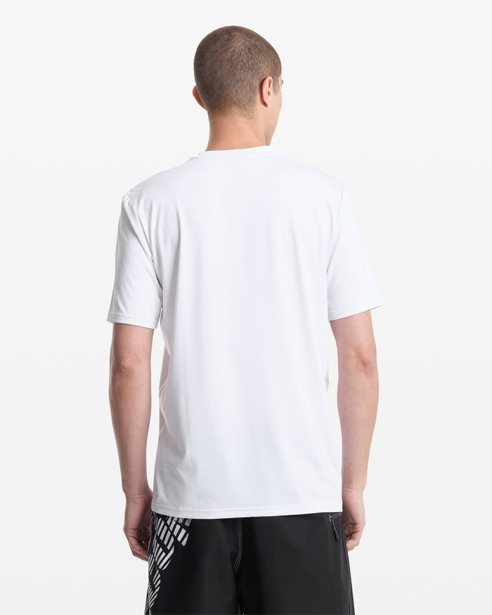 A person with short hair stands facing away from the camera, wearing a plain white Volcom Men's Modstone Loose Short Sleeve Surf Tee and black shorts with a white graphic design on the left side. The background is white.